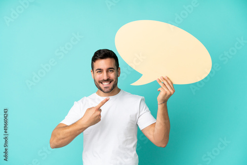 Young handsome caucasian man isolated on blue background holding an empty speech bubble and pointing it