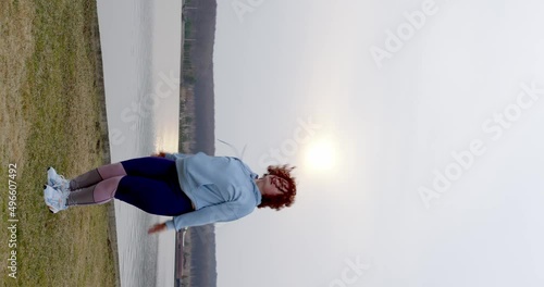 Wallpaper Mural a curly-haired girl with red hair in leggings and a blue bike jumps and simultaneously raises her hands and claps her hands above her head on the lawn against the backdrop of the lake. vertical view Torontodigital.ca
