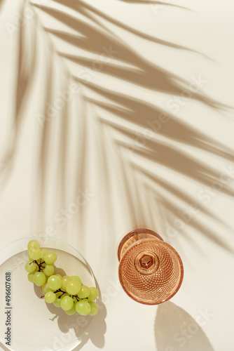 White wine glass peach color glass and fruit grape on beige background, palm leaf shadow at sunlight. Minimal summer alcoholic drink concept. Dry wine in colored glassware, pastel colors