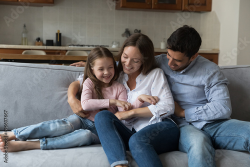 Happy joyful parents, mom and dad cuddling, tickling little daughter kid, sitting, playing on comfortable couch, having fun, enjoying family funny leisure time, parenthood, laughing
