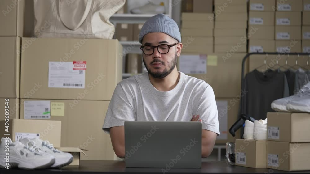 Online seller entrepreneur in a warehouse looking at laptop and worrying about e-commerce business due to lack of sales and customers. Online seller entrepreneur in a warehouse looking at laptop and worrying about e-commerce business due to lack of sales and customers.