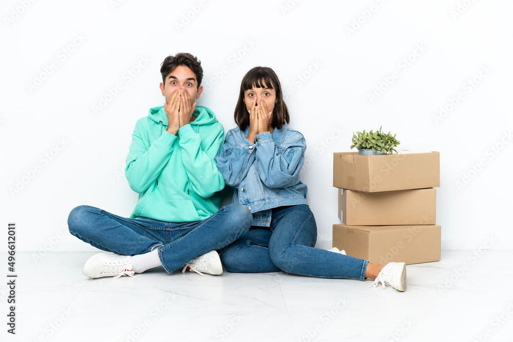 Young couple making a move while picking up a box full of things sitting on the floor isolated on white background smiling a lot while covering mouth Young couple making a move while picking up a box full of things sitting on the floor isolated on white background smiling a lot while covering mouth