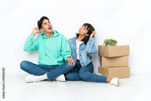 Young couple making a move while picking up a box full of things sitting on the floor isolated on white background thinking an idea while scratching head © luismolinero