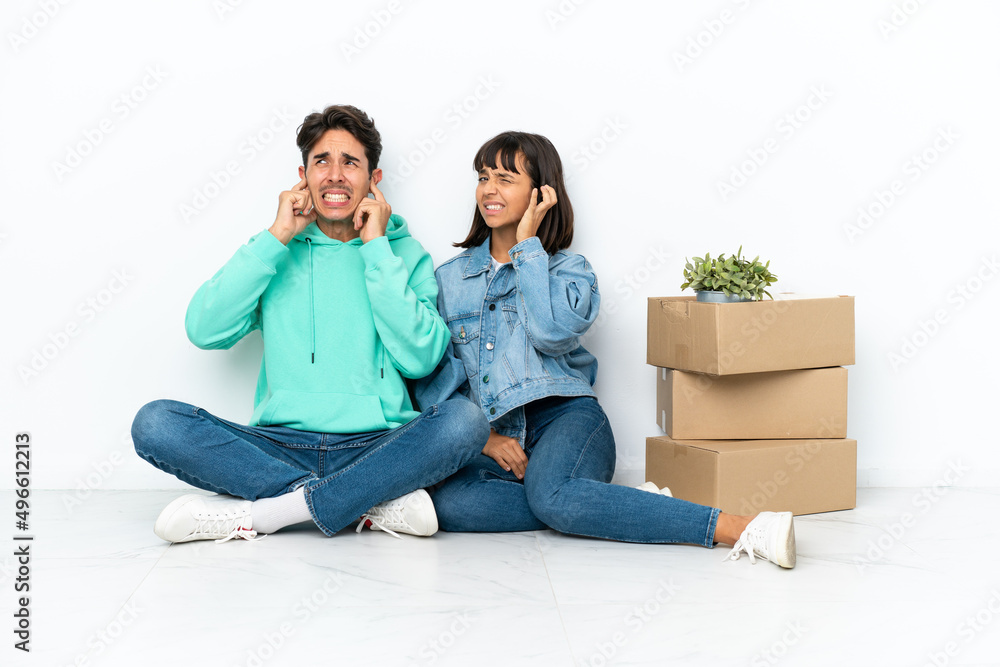 Young couple making a move while picking up a box full of things sitting on the floor isolated on white background covering both ears with hands Young couple making a move while picking up a box full of things sitting on the floor isolated on white background covering both ears with hands
