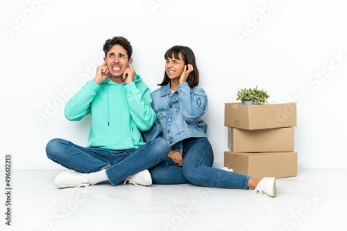 Young couple making a move while picking up a box full of things sitting on the floor isolated on white background covering both ears with hands © luismolinero