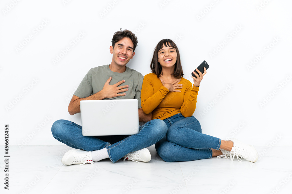 Young couple with a laptop and mobile sitting on the floor smiling a lot while putting hands on chest