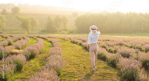 Beautiful young healthy woman with a white dress running joyfully through a lavender field holding a straw hat under the rays of the setting sun