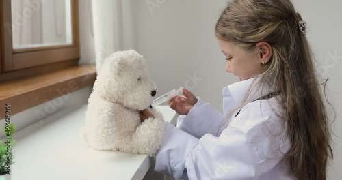 Little girl wear white coat play pediatrician, pretend be doctor or nurse treats fluffy bear holds toy syringe, make injection or vaccine, have fun at home. Healthcare, medicine, vet clinic ad concept photo