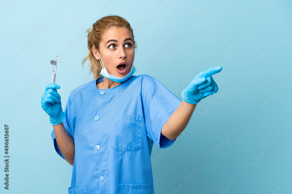 Young woman dentist holding tools over isolated blue background pointing away Young woman dentist holding tools over isolated blue background pointing away