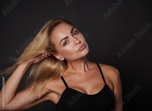 Close up fashion portrait of beautiful European woman with toned body, fresh clean tanned skin touching her shining blond hair and looking at camera on black background