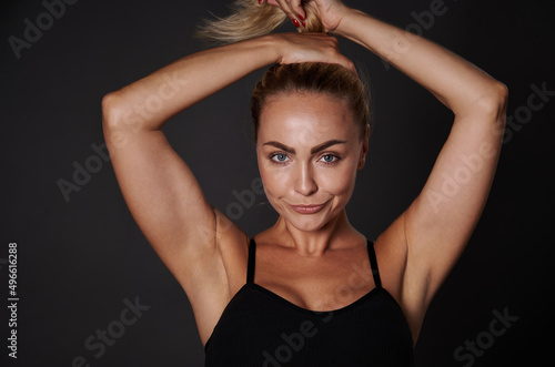 Sexy middle aged woman with perfect body, beautiful healthy tanned skin, posing in black lingerie and tying a ponytail on her head, three quarter studio portrait over dark background with copy space