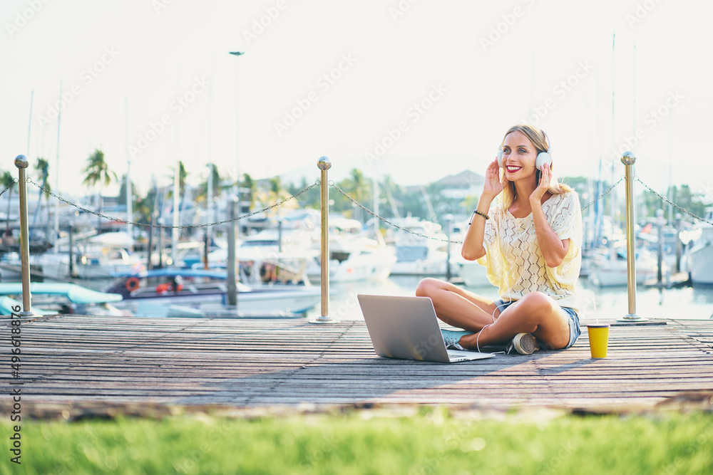 Listening to music and working outdoors. Pretty young woman in headphones using laptop computer at sea marine.