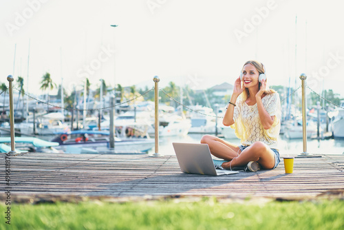 Listening to music and working outdoors. Pretty young woman in headphones using laptop computer at sea marine.