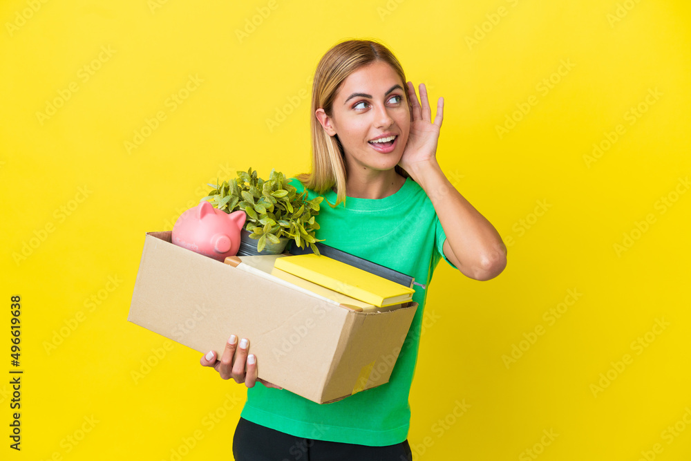 Young Uruguayan girl making a move while picking up a box full of things isolated on yellow background listening to something by putting hand on the ear Young Uruguayan girl making a move while picking up a box full of things isolated on yellow background listening to something by putting hand on the ear