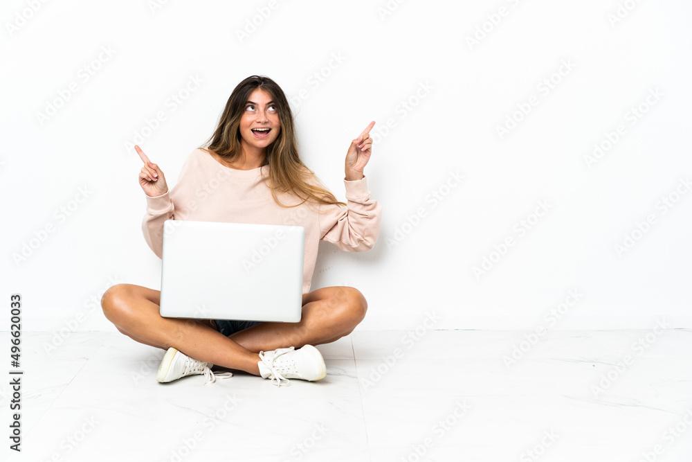 Young woman with a laptop sitting on the floor isolated on white background pointing finger to the laterals and happy