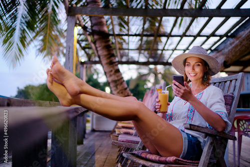 Vacation and technology. Young pretty woman in hat using smartphone sitting at beach cafe bar.