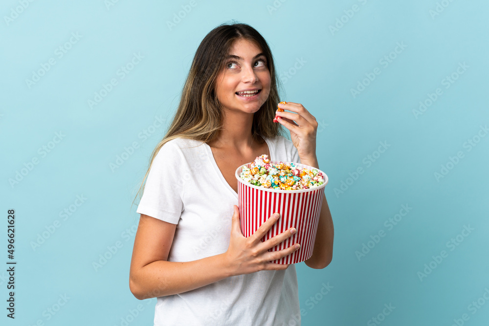 Young caucasian woman isolated on blue background holding a big bucket of popcorns