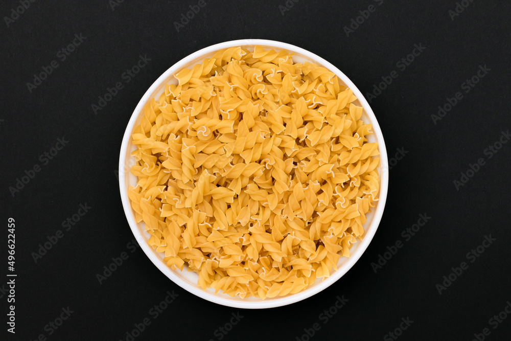 Uncooked Fusilli Pasta Lying on White Plate on Black Background. Raw and Dry Macaroni. Unhealthy and Fat Food - Top View