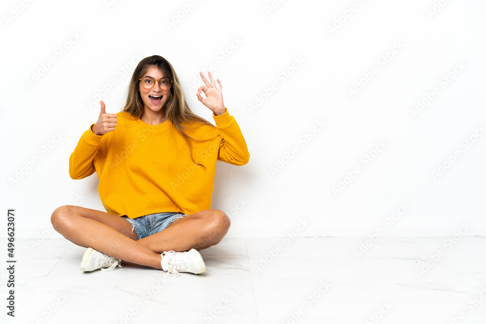 Young woman sitting on the floor isolated on white background showing ok sign and thumb up gesture Young woman sitting on the floor isolated on white background showing ok sign and thumb up gesture