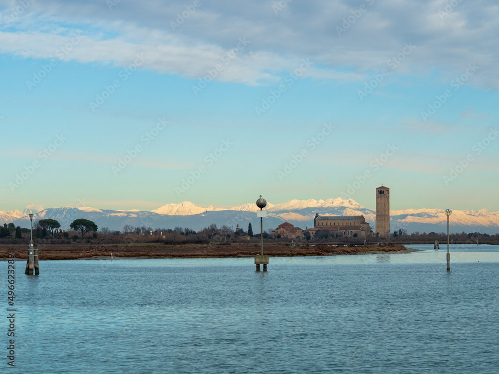 View from Burano across the water towards the island of Torcello and the church of Basilica di Santa Maria Assunta with the snow-capped Dolomite mountains in the background - Torcello, Venice, Italy