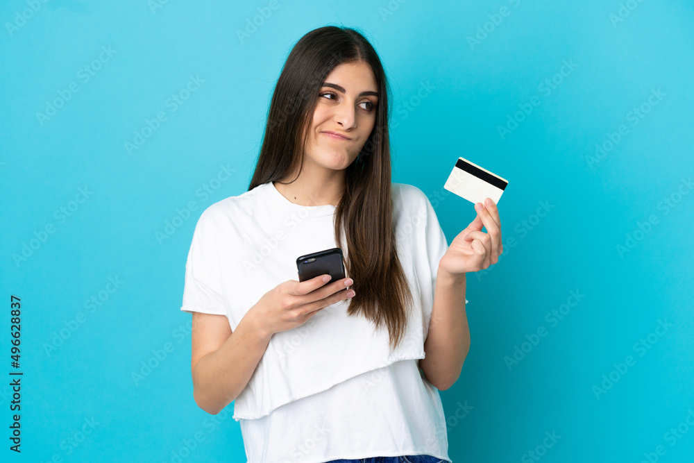 Young caucasian woman isolated on blue background buying with the mobile with a credit card while thinking Young caucasian woman isolated on blue background buying with the mobile with a credit card while thinking