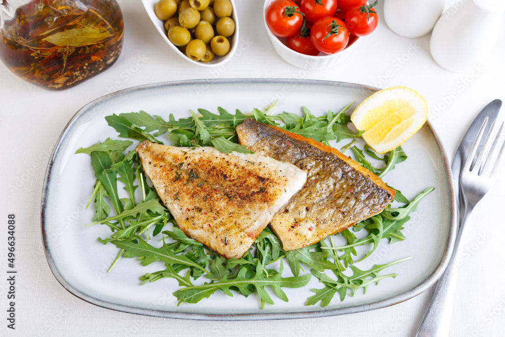 Fried dorado fillet with crispy skin. Arugula, cherry tomatoes, olives, lemon and olive oil. Traditional Mediterranean dish. White background. Selective focus, close-up, top view. Fried dorado fillet with crispy skin. Arugula, cherry tomatoes, olives, lemon and olive oil. Traditional Mediterranean dish. White background. Selective focus, close-up, top view.
