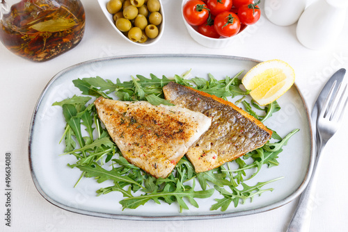 Fried dorado fillet with crispy skin. Arugula, cherry tomatoes, olives, lemon and olive oil. Traditional Mediterranean dish. White background. Selective focus, close-up, top view.