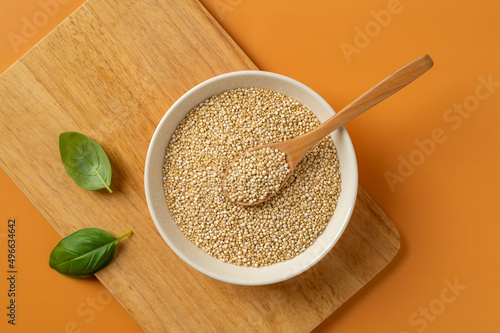 White quinoa grains and spoon in a beige bowl on a wooden cutting board over orange background. Chenopodium quinoa seeds for gluten free dieting. Concepts of superfood vegetarian healthy eating.