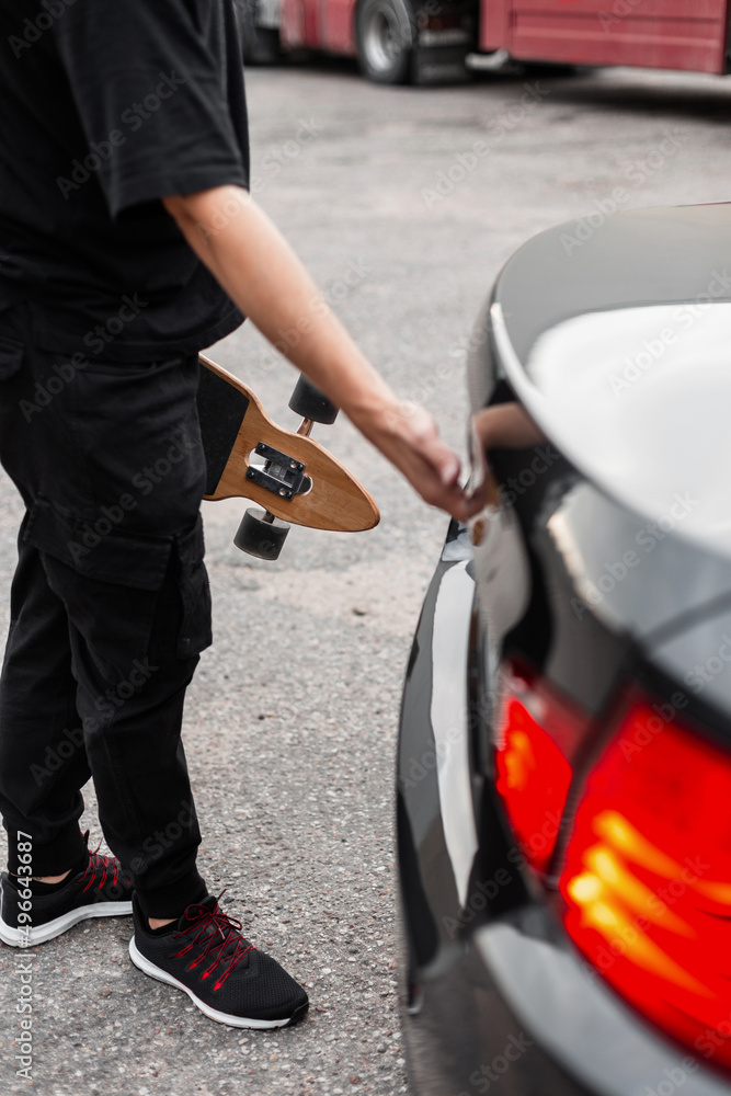 Young man in black clothes with longboarding opens the trunk of a black car. Active weekend concept