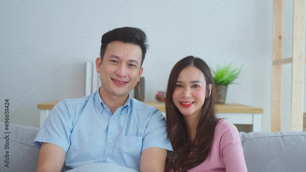 Young happy asian couple looking at camera smiling and laughing sitting together on sofa couch in living room at home, healthy relationship, married life. Young happy asian couple looking at camera smiling and laughing sitting together on sofa couch in living room at home, healthy relationship, married life.