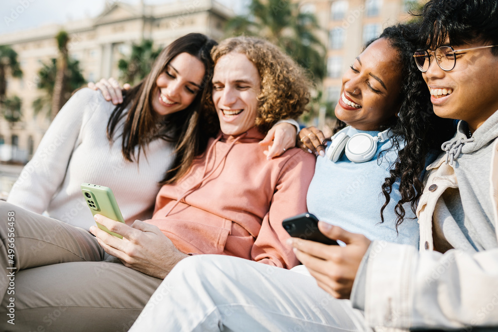 Group of multiracial young trendy friends using cell phones sharing content on social media platform app - United millennial people holding smartphones sitting outdoor in city street