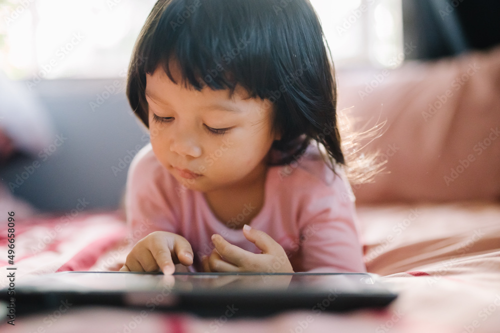 A little girl in a pink shirt lay intently playing the tablet on her bed. self-learning technology education concept