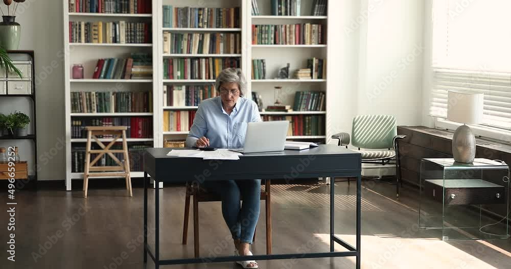 In office middle-aged 60s woman in glasses pay bills, working sit at desk use laptop, make payments via e-bank app. Company finances control, money management, accountancy job use modern tech concept In office middle-aged 60s woman in glasses pay bills, working sit at desk use laptop, make payments via e-bank app. Company finances control, money management, accountancy job use modern tech concept