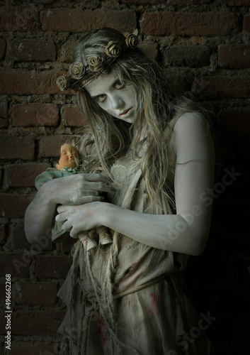 A girl is dressed in rags as a Halloween horror 
figure. She stands in front of an old brick wall. 
Her clothes are torn and dirty and she holds a 
broken doll in her arms.
 photo