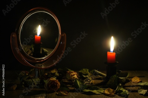 Картината върху платно A burning candle reflected in a mirror standing on a wooden table among withered roses