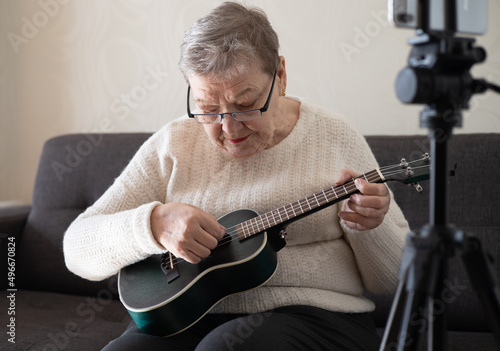 Elderly woman 70+ sits on the couch and learns to play the ukulele at home using video tutorial on her smartphone. Adaptation of pensioners in the modern world. New knowledge and skills. Hobby
