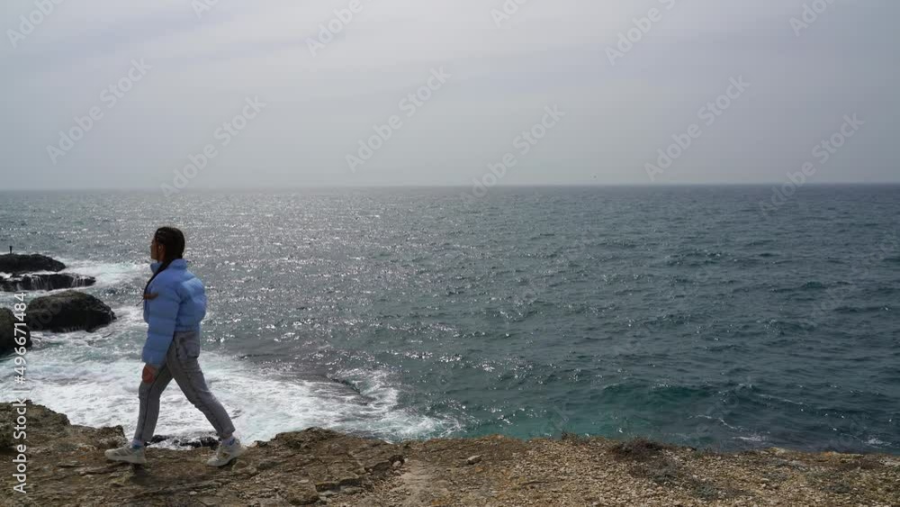 A woman in a blue jacket walks on a cliff above a cliff overlooking the sea, looking at the stormy ocean. Girl traveler rests, thinks, dreams, enjoys nature. Peace and calm landscape, windy weather. A woman in a blue jacket walks on a cliff above a cliff overlooking the sea, looking at the stormy ocean. Girl traveler rests, thinks, dreams, enjoys nature. Peace and calm landscape, windy weather.
