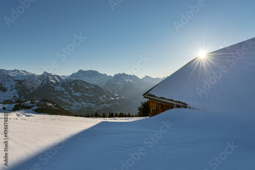 Verschneite Bergwiesen mit Heuhütten vor Panorama der Glarner Alpen mit der Abendsonne, Kanton Sankt Gallen, Schweiz