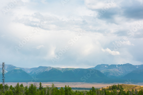 Dramatic view from forest to high mountain range in sunlight during rain in changeable weather. Colorful landscape with green forest and sunlit steppe against large mountains under cloudy sky in rain.
