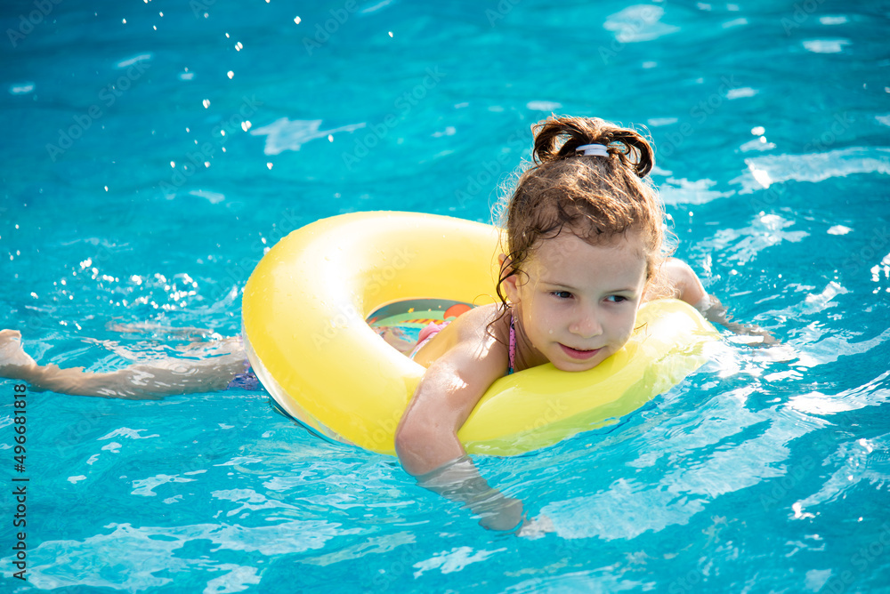 A beautiful little smiling girl in a swimsuit and in a yellow inflatable circle bathes and plays in the pool with clear blue water. Summer. Vacation. Rest.