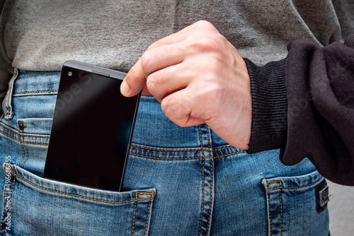 The hand of a thief pulling smartphone out of someone's pocket. Blue jeans pocket with gadget. Closeup concept of theft, crime, dishonesty