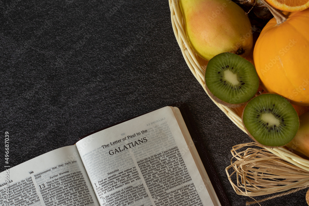 An open Holy Bible book of Galatians with a wicker basket full of various fresh fruits with copy