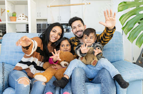 Photography Happy Indian couple and their young children waving hands looking at camera sitting on sofa at home