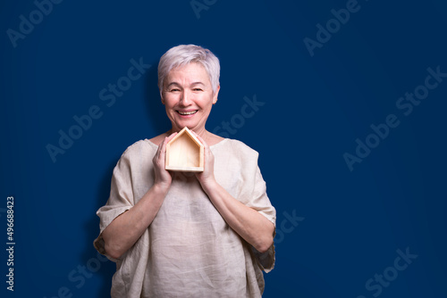 A beautiful elderly woman with a wooden house in her hands. Investing savings. Buying a home. Copy space. Blue background.