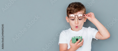 Shocked child boy using smartphone and taking off glasses on gray background