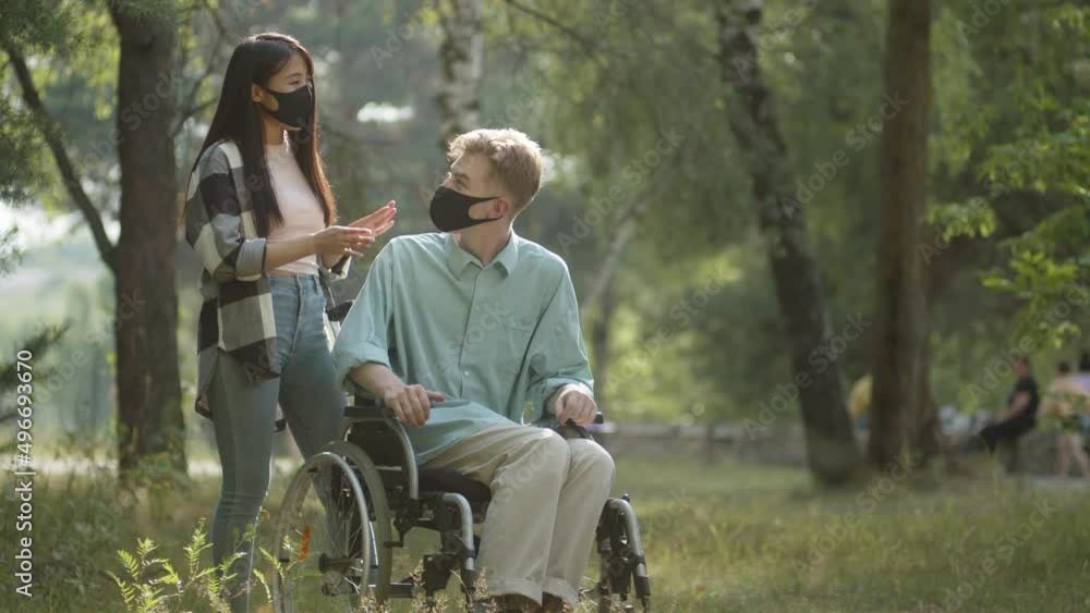 Being in the public park, a handsome asian lady rolls her disabled blonde man in a wheelchair, then they stop, talk a little and decide move away Being in the public park, a handsome asian lady rolls her disabled blonde man in a wheelchair, then they stop, talk a little and decide move away