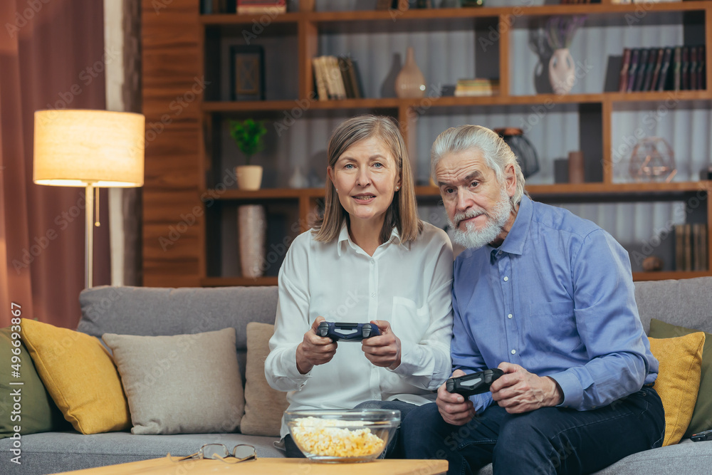 senior couple playing video game console with joysticks sitting on couch in living room. Funny Mature people family having fun spend leisure time together. gray hair man and woman enjoy entertaining senior couple playing video game console with joysticks sitting on couch in living room. Funny Mature people family having fun spend leisure time together. gray hair man and woman enjoy entertaining