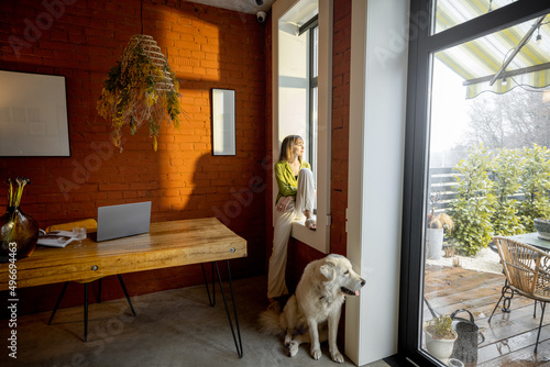 Woman sitting on the window sill and looking out the window with her white adorable dog at home. Wide view on interior and backyard