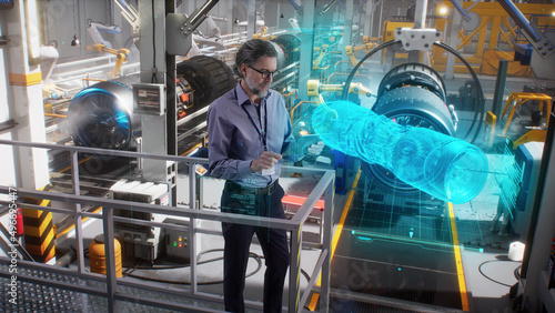 An engineer uses a tablet with a holographic innovative application, to model and design and monitor a 3D model of an aircraft engine while on an automated robotic line at an aerospace factory © Framestock