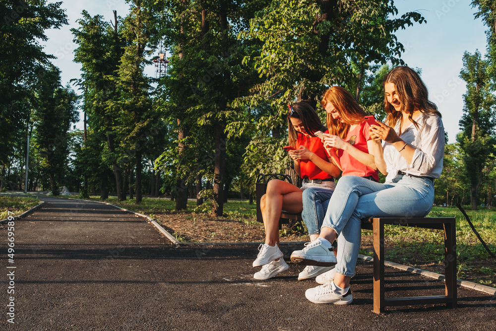 Diverse three girl friends using their phones outdoors. Group gen z young people using mobile smartphone sitting on bench in summer park